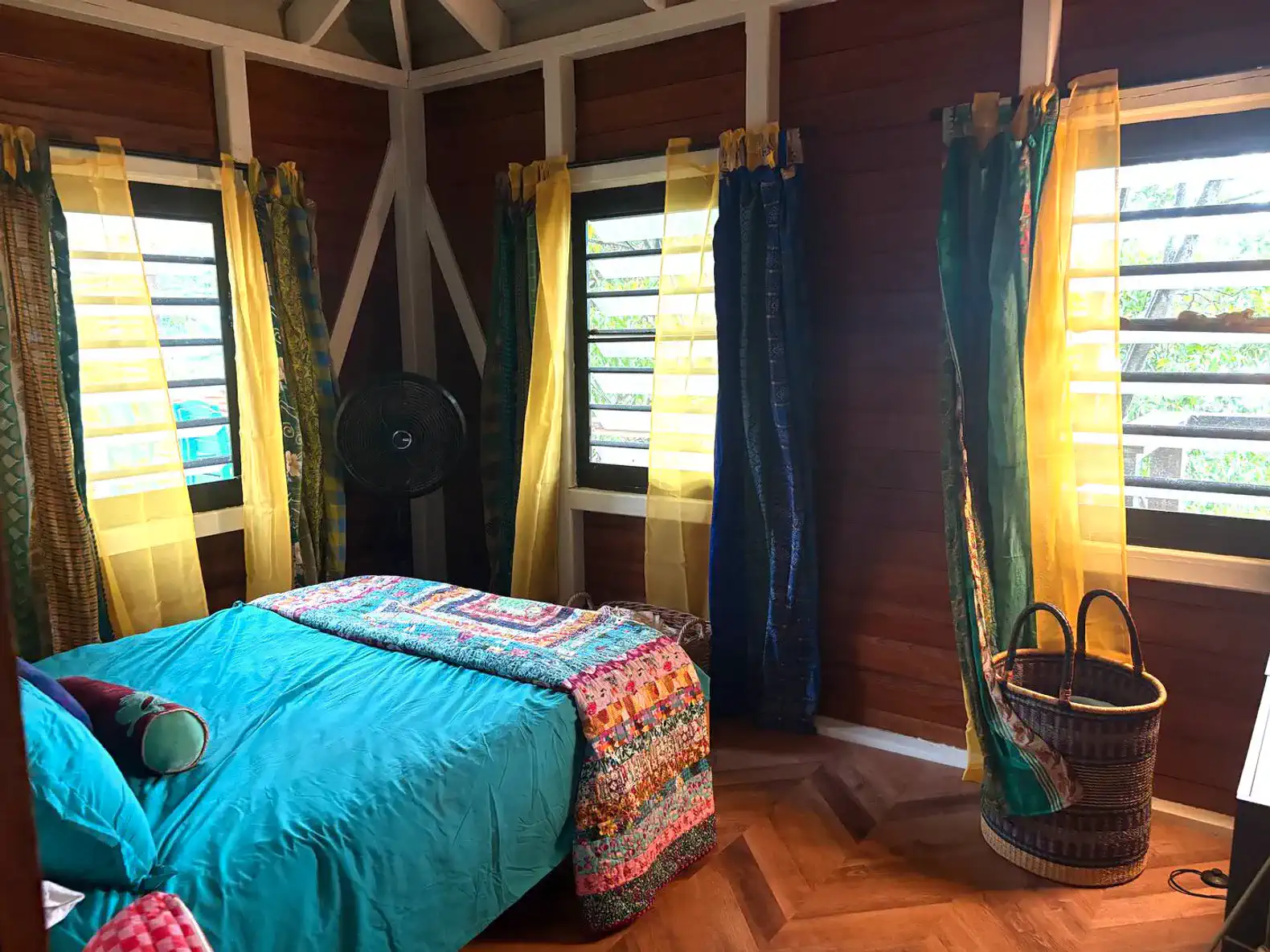 Brightly colored bedroom with a turquoise bedspread, patterned throw, and sheer yellow curtains letting in natural light