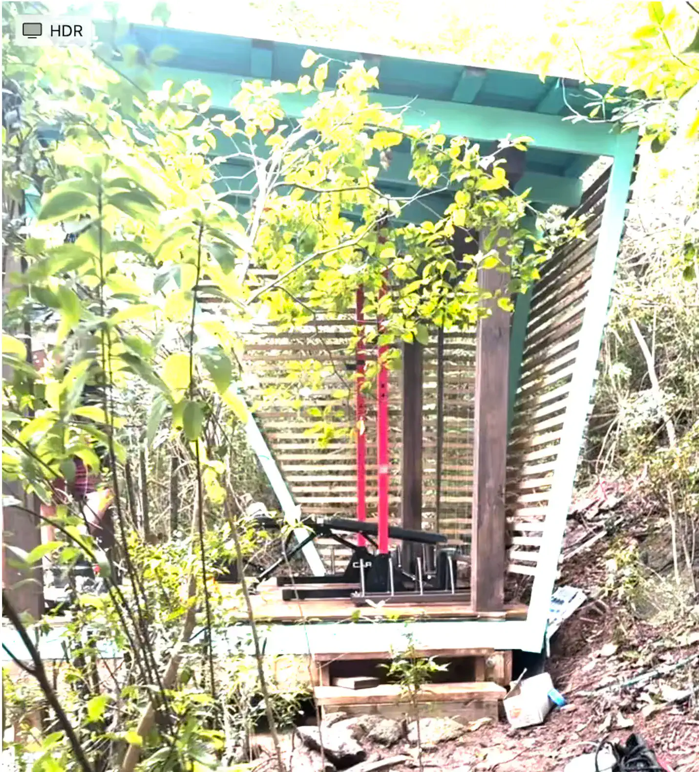 Outdoor fitness area surrounded by lush greenery, featuring exercise equipment under a slatted roof