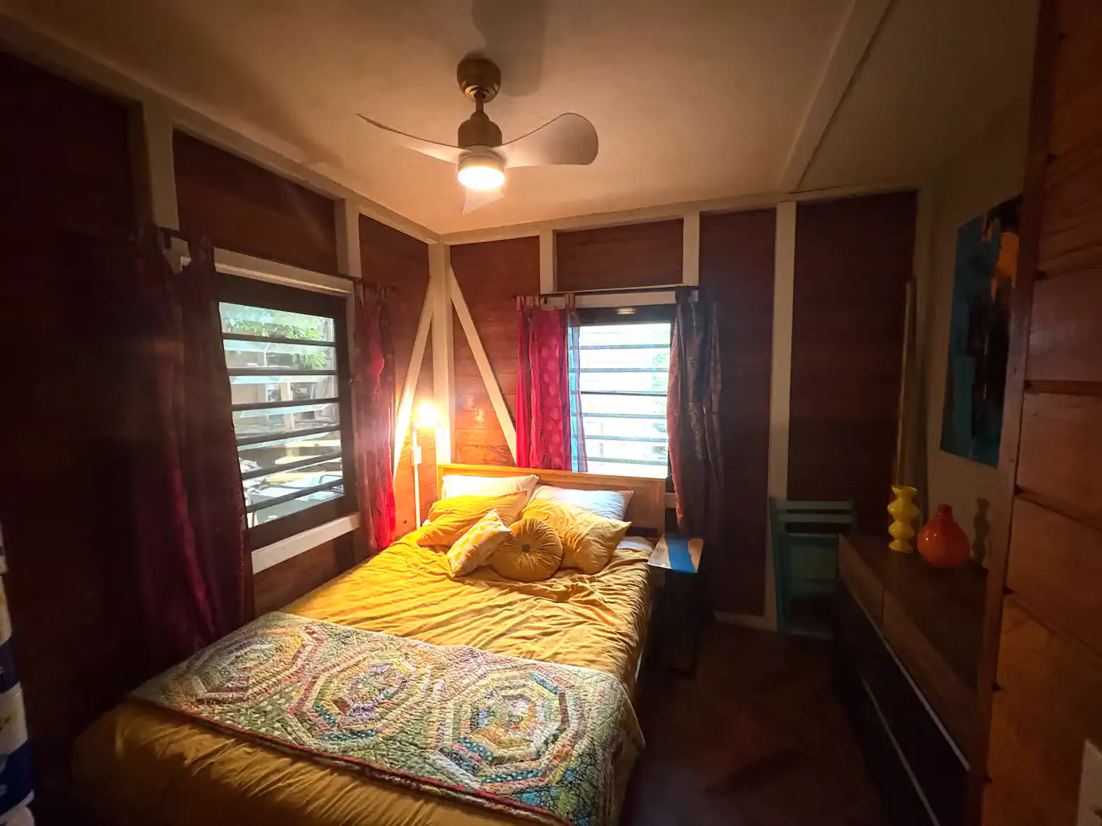 Cozy bedroom with a queen bed, colorful bedding, and wooden accents, featuring a ceiling fan and bright curtains