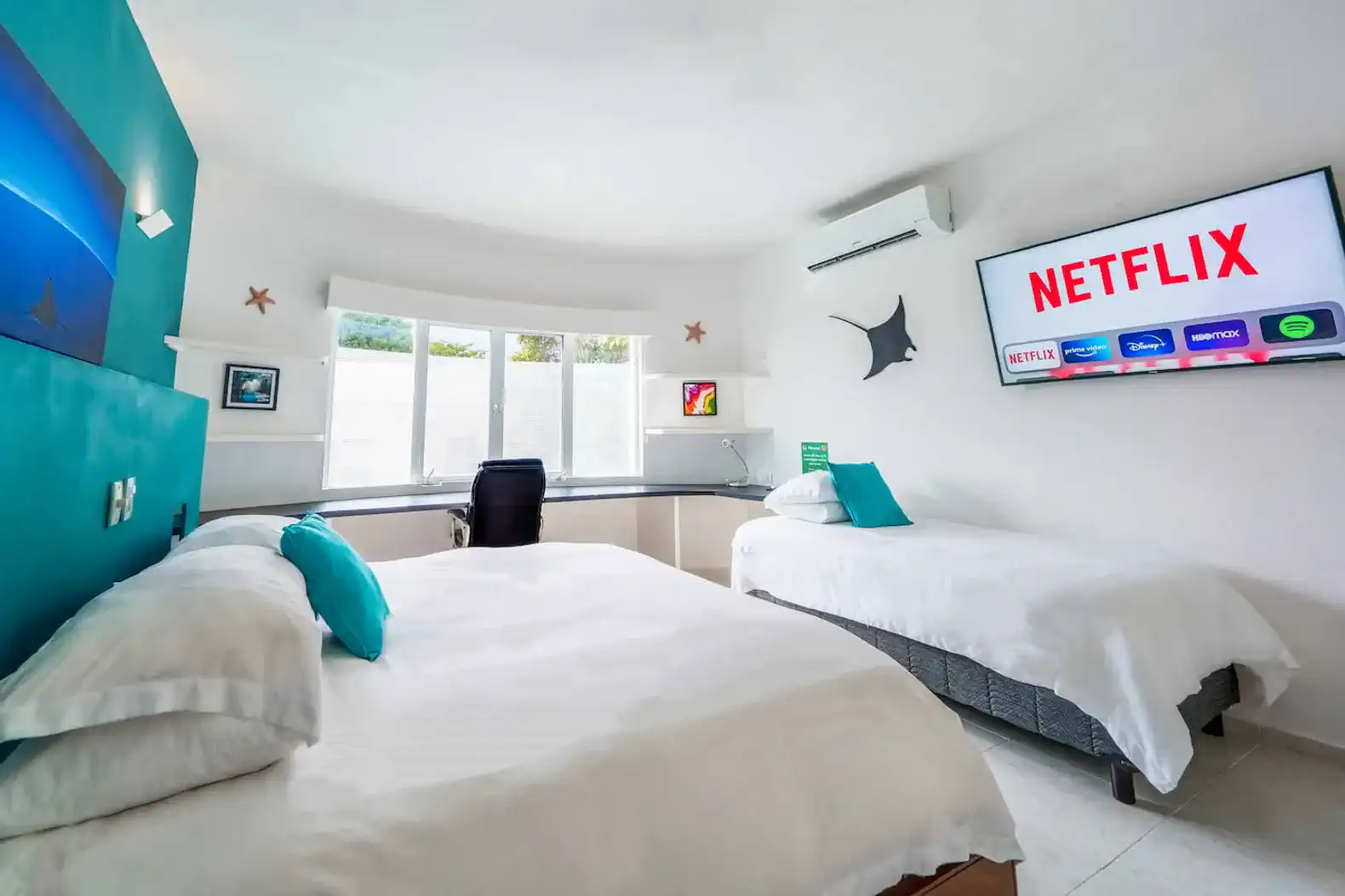 Bright guest bedroom with two beds, large windows, and a wall-mounted TV