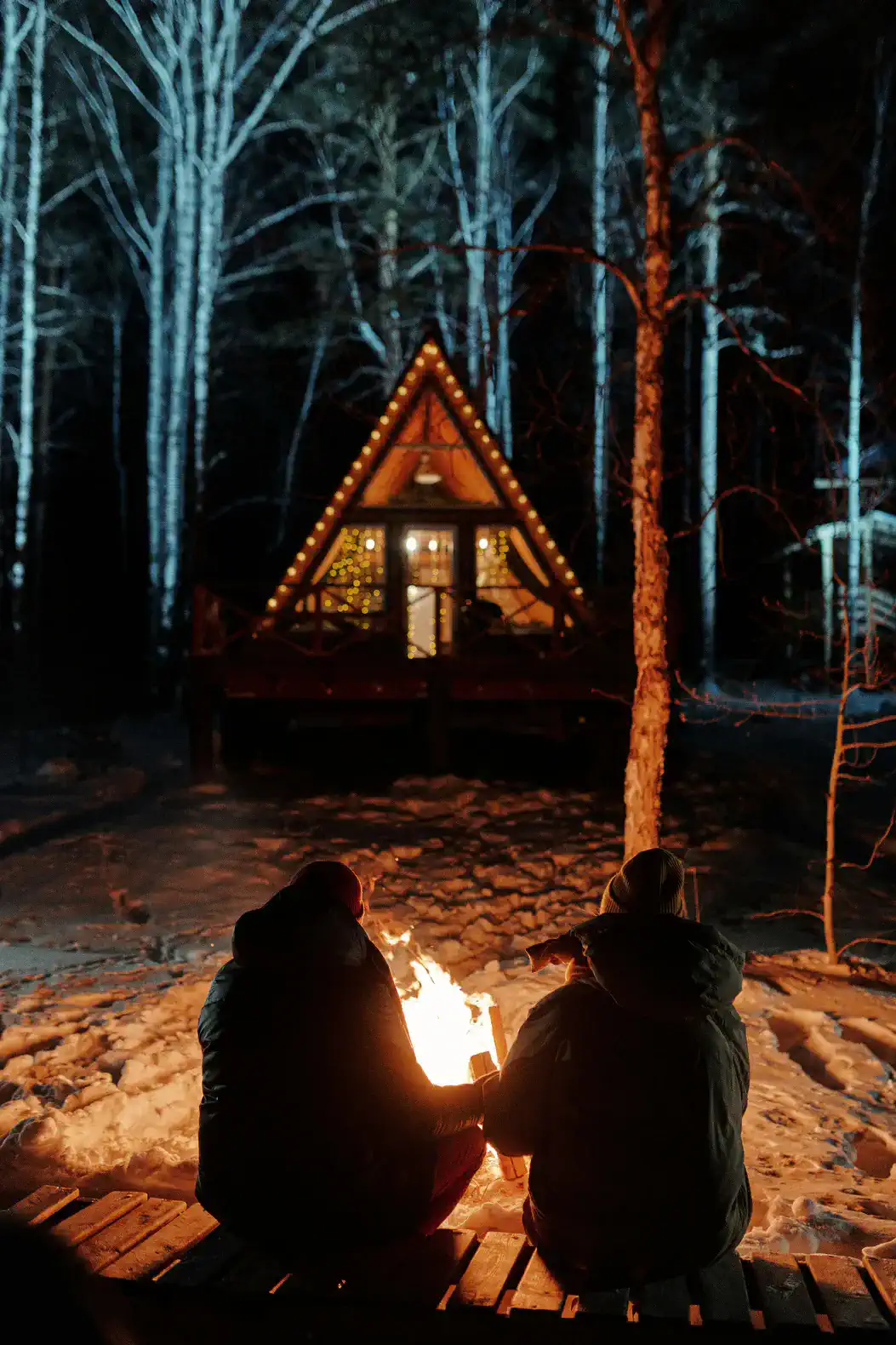 Cozy outdoor fire pit area with two people sitting by the flames in a snowy forest setting