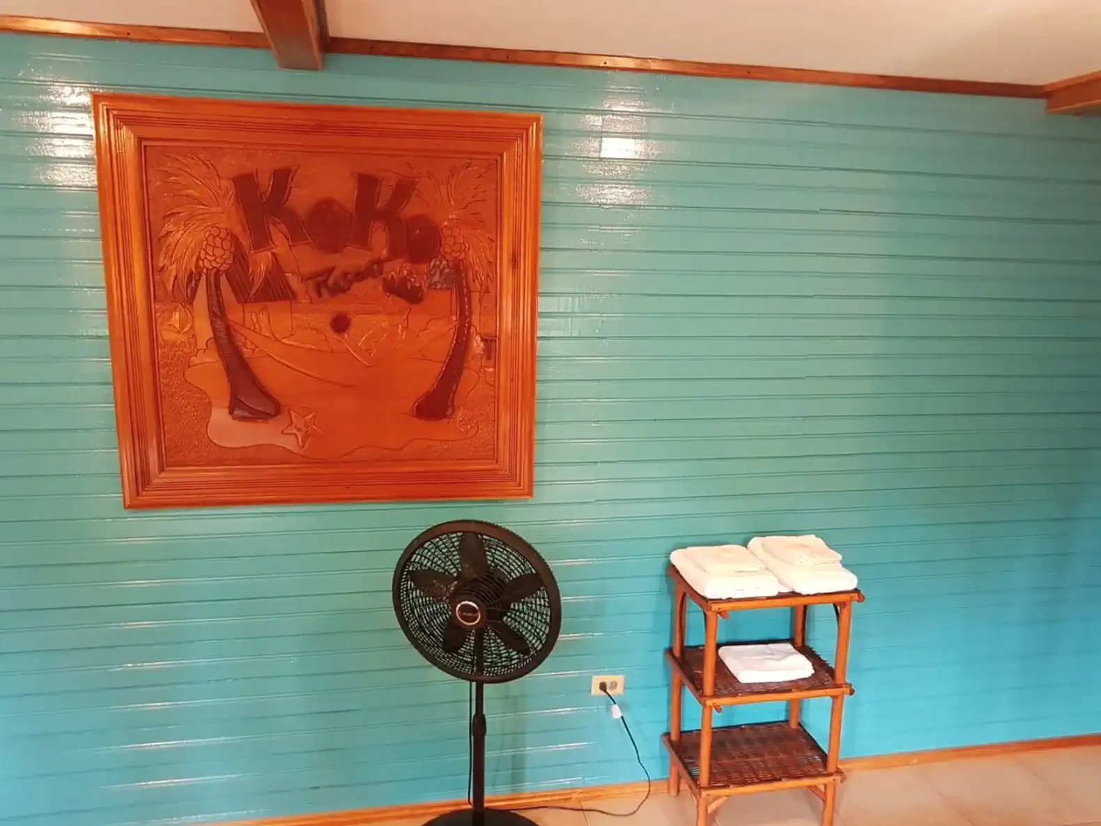 Brightly colored wall with a wooden artwork and a fan beside a shelf of towels