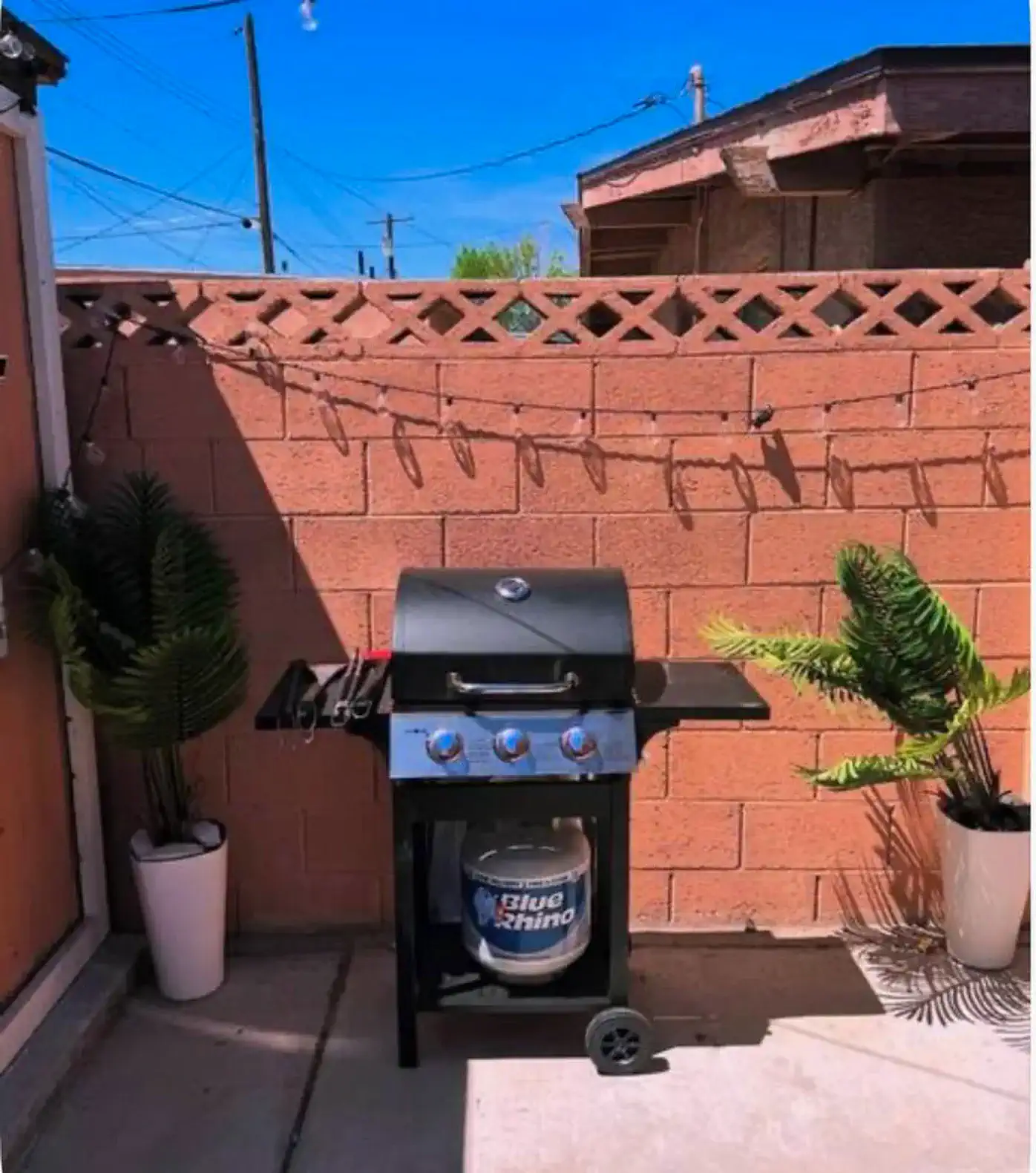 Outdoor BBQ grill with potted plants and string lights against a brick wall