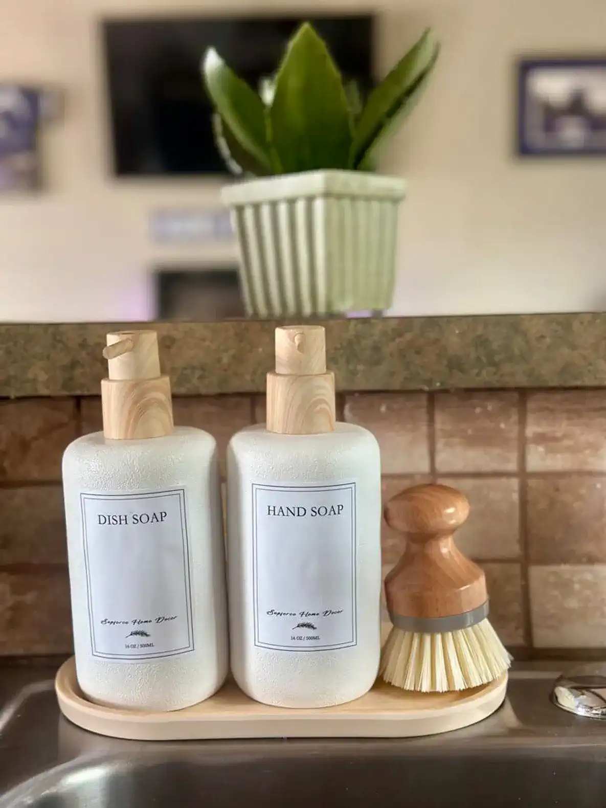 Stylish soap dispensers and a brush on a kitchen counter with a plant in the background