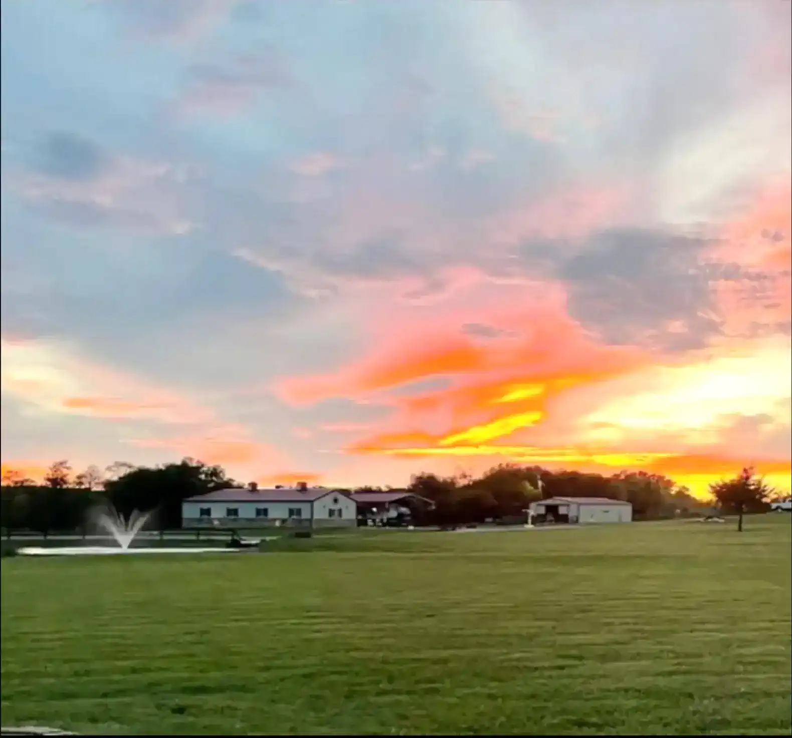 Colorful sunset over a grassy area with a fountain and buildings in the background