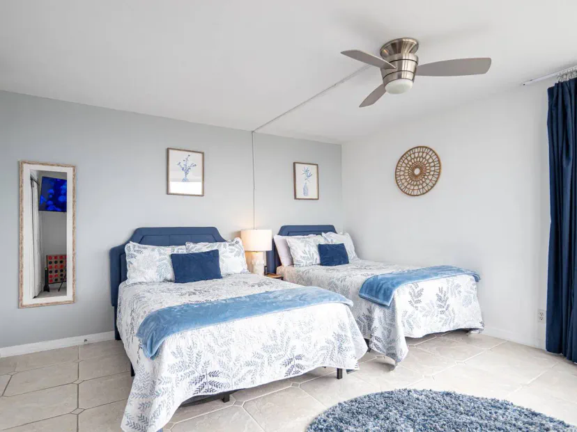 Spacious bedroom with two double beds, blue accents, and a modern ceiling fan, featuring natural light and tiled flooring