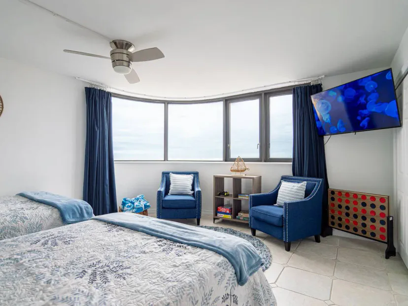 Dormitorio acogedor con dos camas, sillones azules, grandes ventanas y un televisor, con vistas al océano y decoración de playa