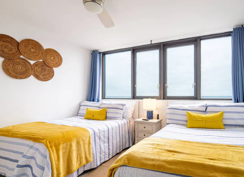 Dormitorio luminoso con dos camas, decoración costera, grandes ventanas que ofrecen vistas al océano y suaves acentos amarillos
