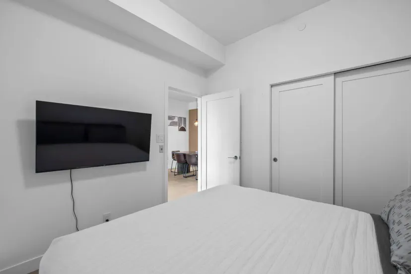 Cozy bedroom with a queen bed, wall-mounted TV, and closet space, leading to an open living area