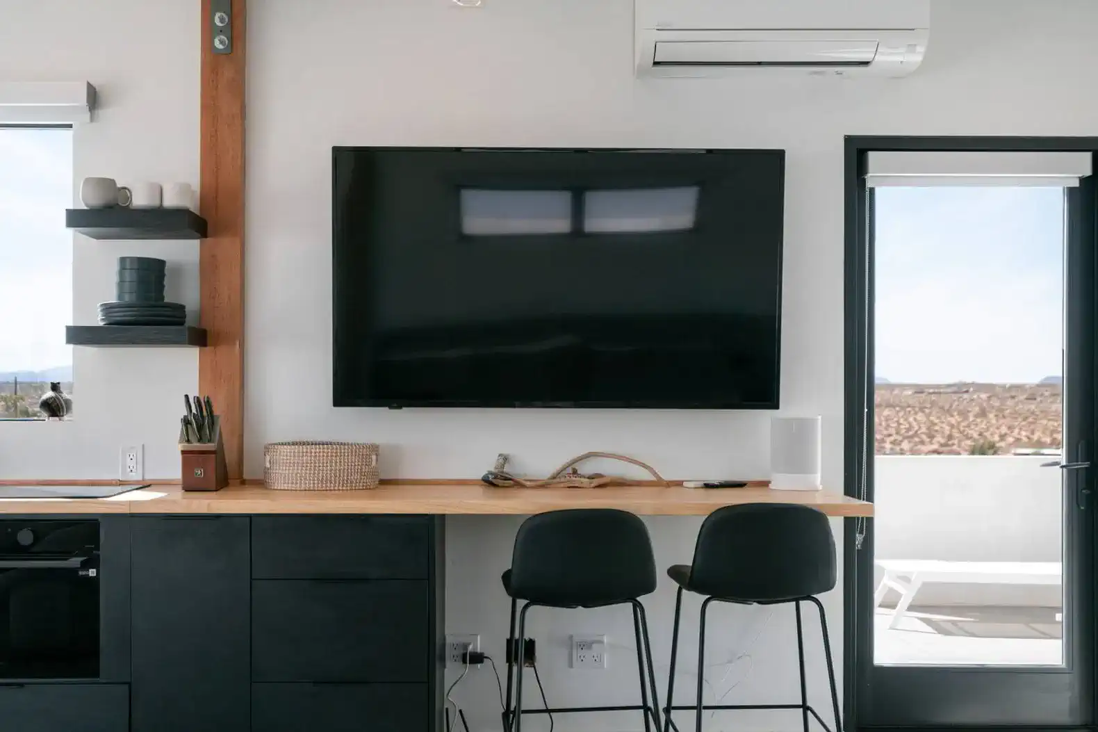 Modern kitchen area with a large TV, bar stools, and a view of the desert outside