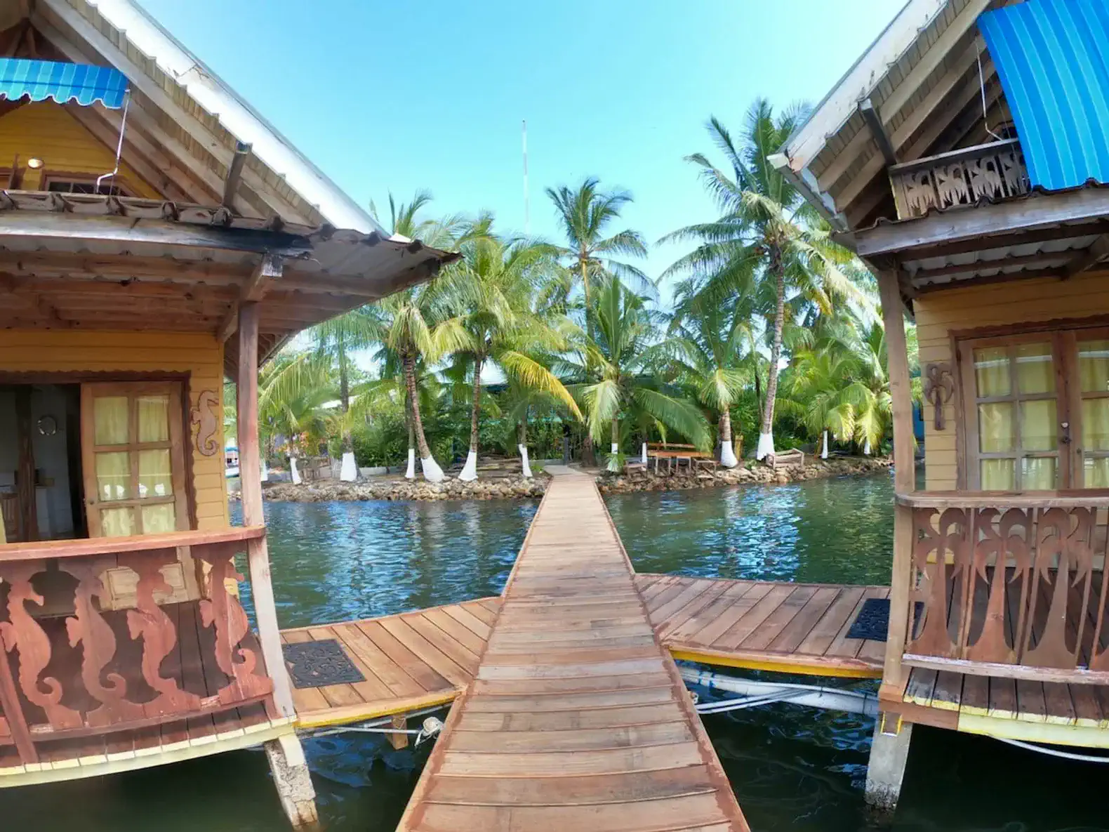 Wooden walkway connecting two bungalows surrounded by palm trees and water