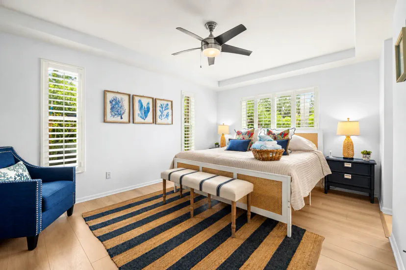 A spacious and inviting primary bedroom blends relaxed coastal style with plenty of natural light, featuring a king-size bed, comfortable sitting area, plantation shutters and warm wood flooring. Thoughtful blue accents and fresh linens create a serene ret