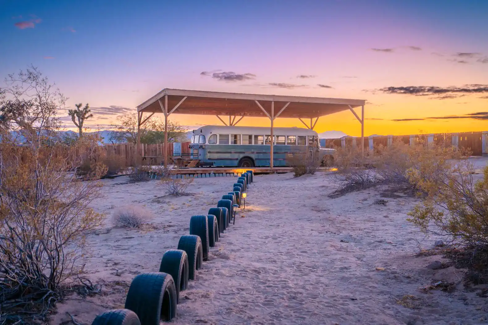 Rustic bus under a wooden shelter at sunset with desert landscape and tire path