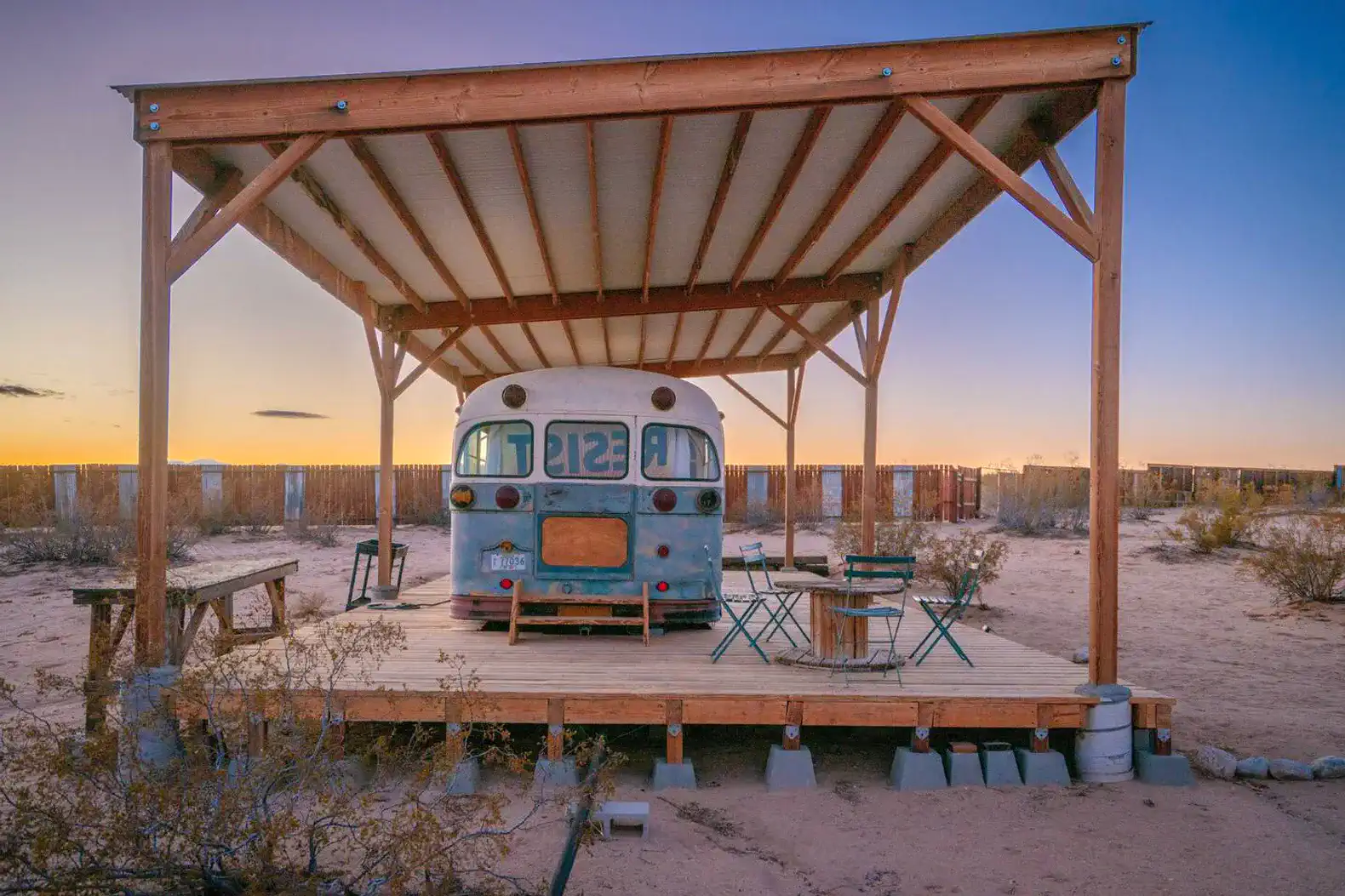 Rustic bus under a wooden shelter in a desert setting with outdoor seating