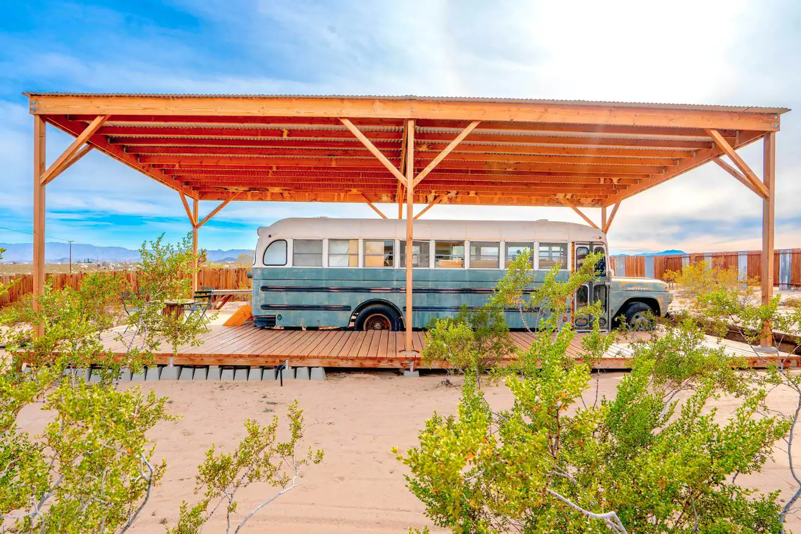 Converted bus under a wooden shelter surrounded by desert landscape