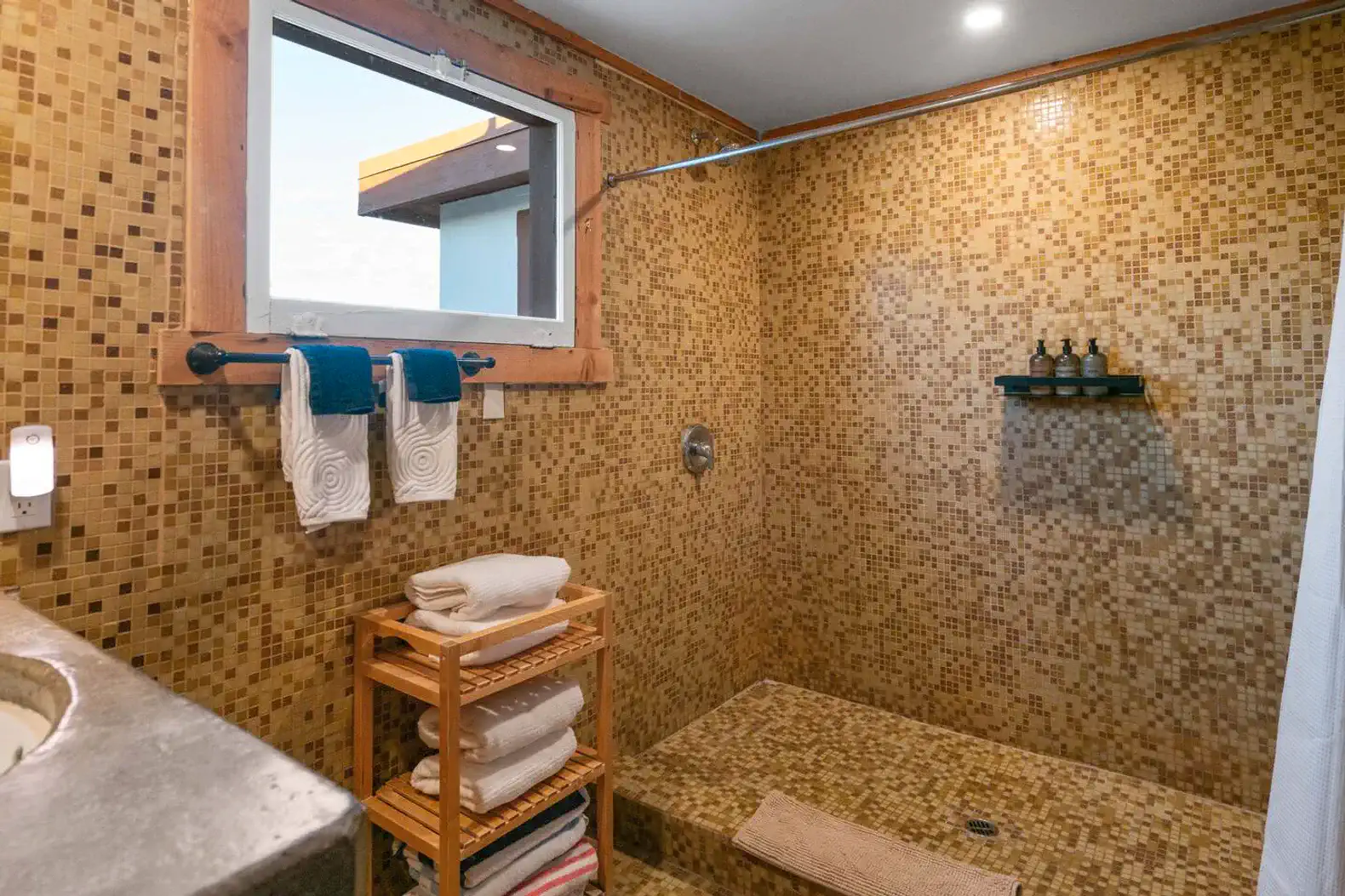 Spacious shower with mosaic tiles and wooden accents, featuring towels and a shelf