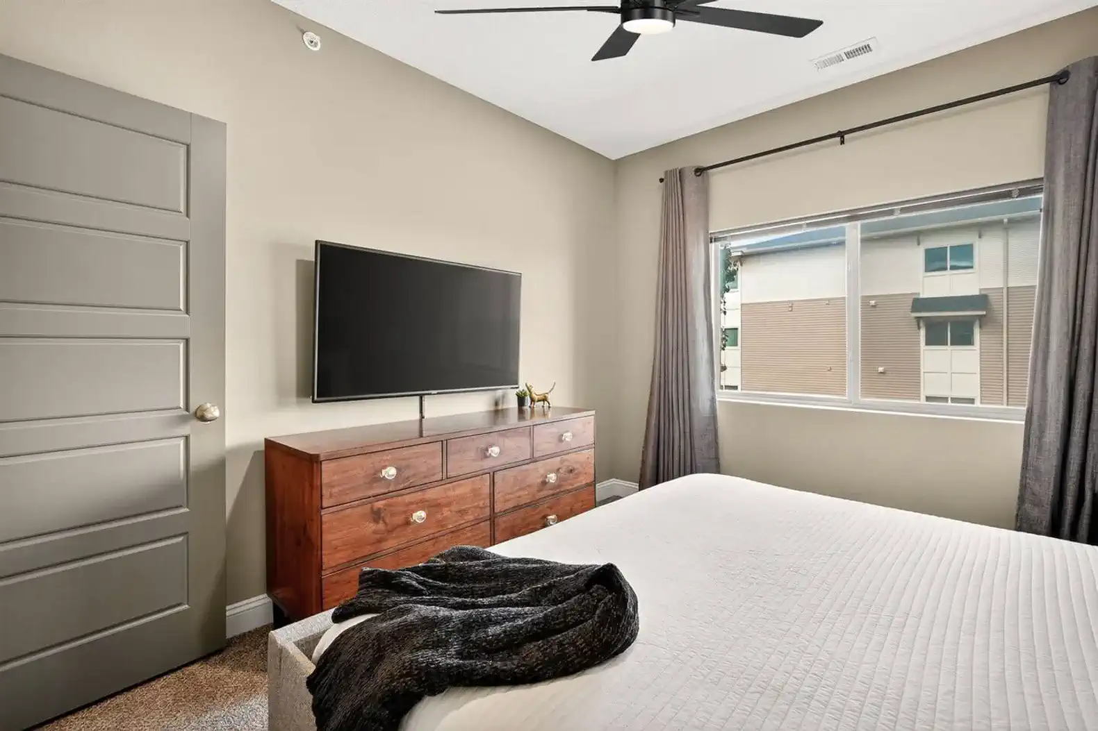 Bedroom retreat featuring a 55" smart TV, sleek dresser, blackout curtains, ceiling fan and stylish décor for a condo-inspired stay.