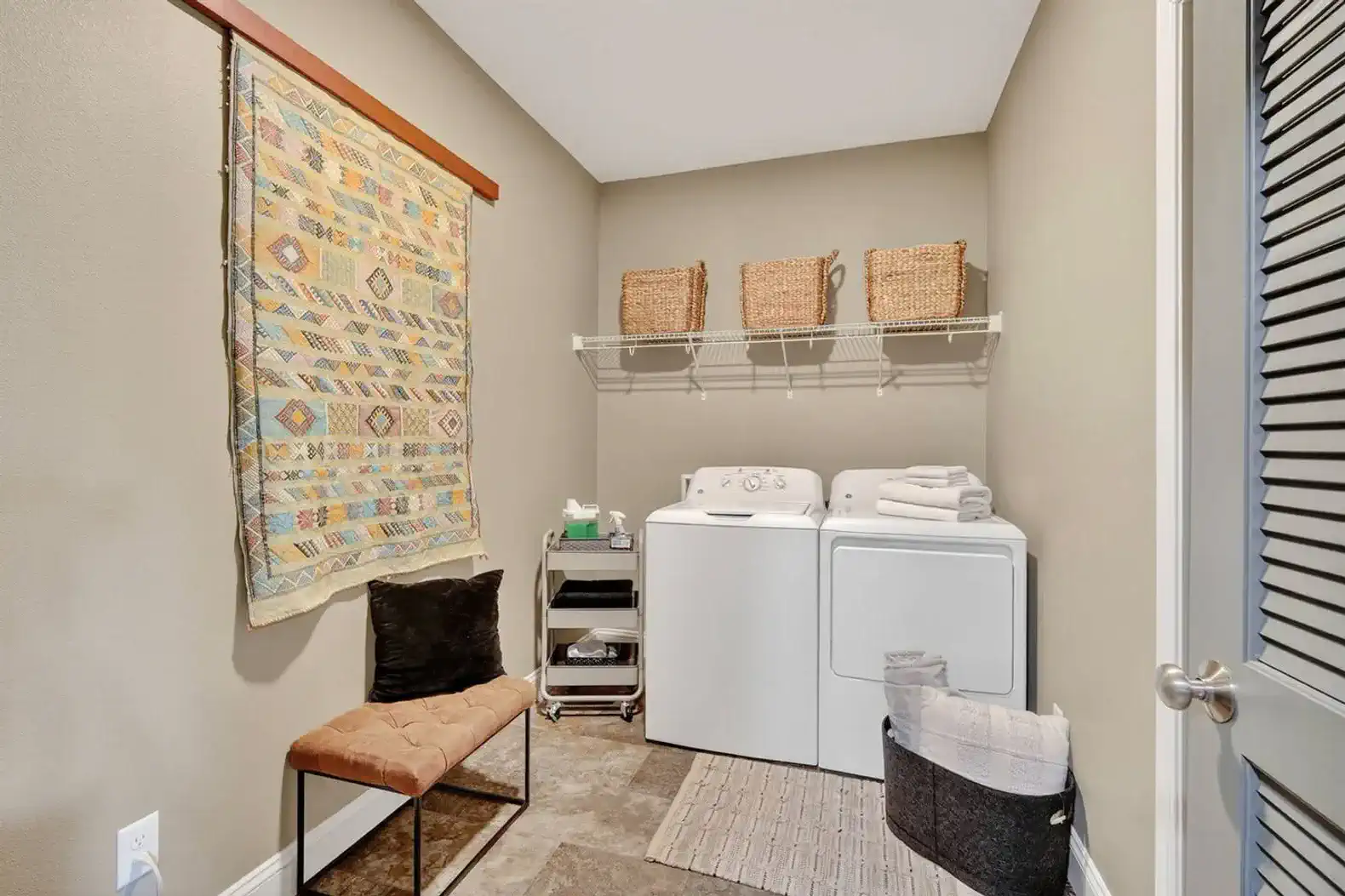 Convenience meets style in the oversized laundry room with washer, dryer, and open shelving. Thoughtful touches like a decorative wall hanging and bench seating elevate this space.