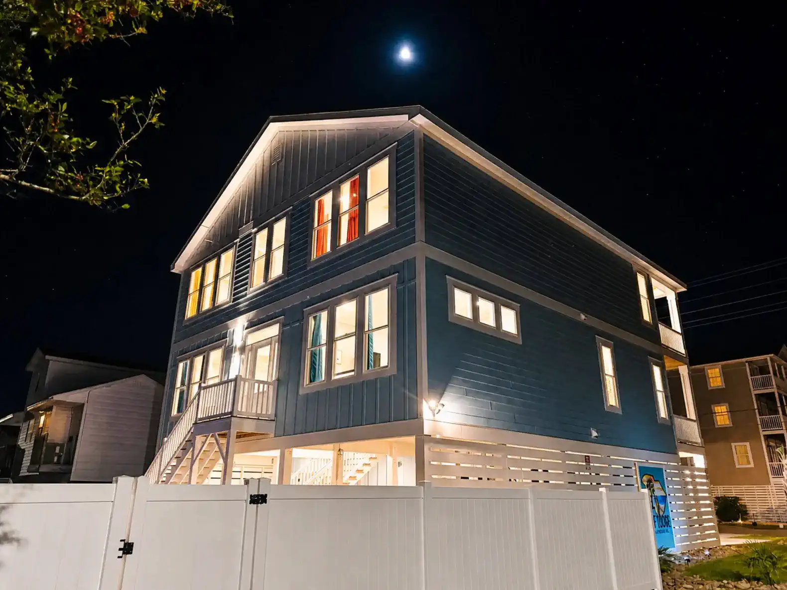 Exterior view of a large blue house at night with illuminated windows and a white fence