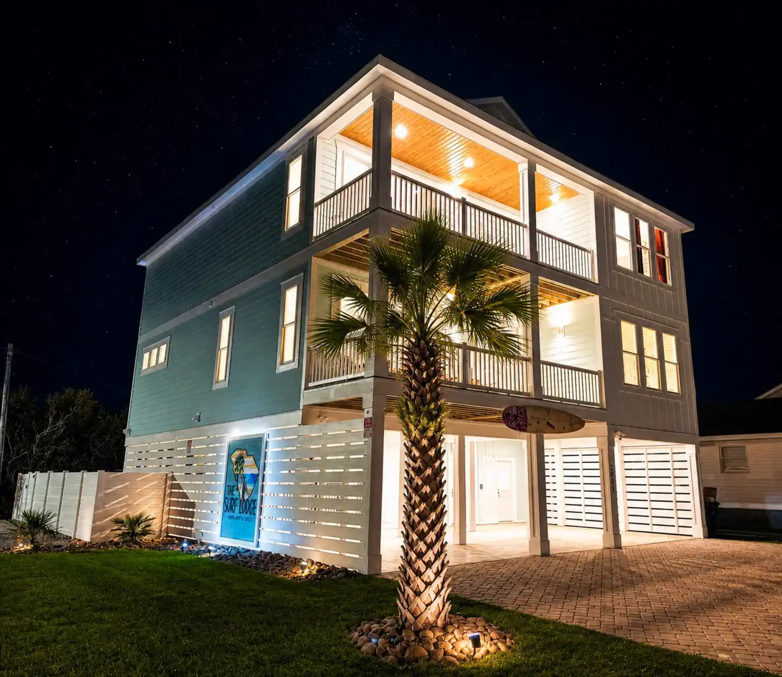 Exterior view of a large beach house with palm trees and illuminated balconies at night
