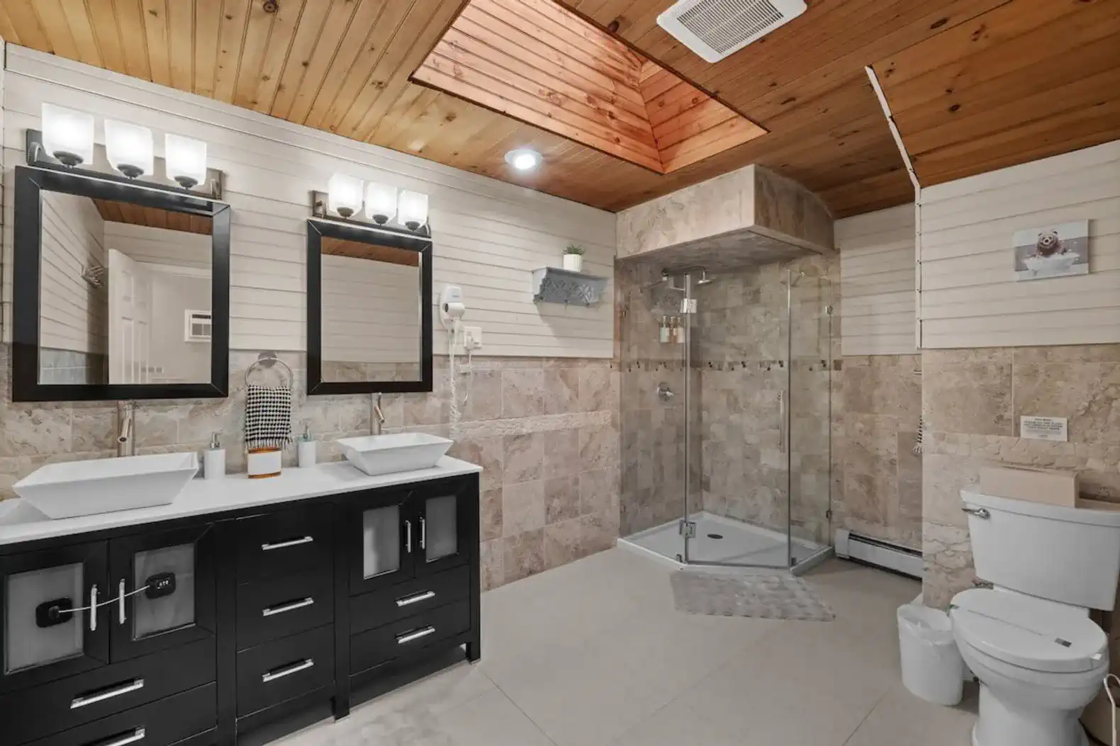 Luxurious full bathroom with double vanity sinks, walk-in glass shower, and elegant stone tile finishes. Features a wood-accent ceiling, bright lighting, and added comforts like a built-in hair dryer.