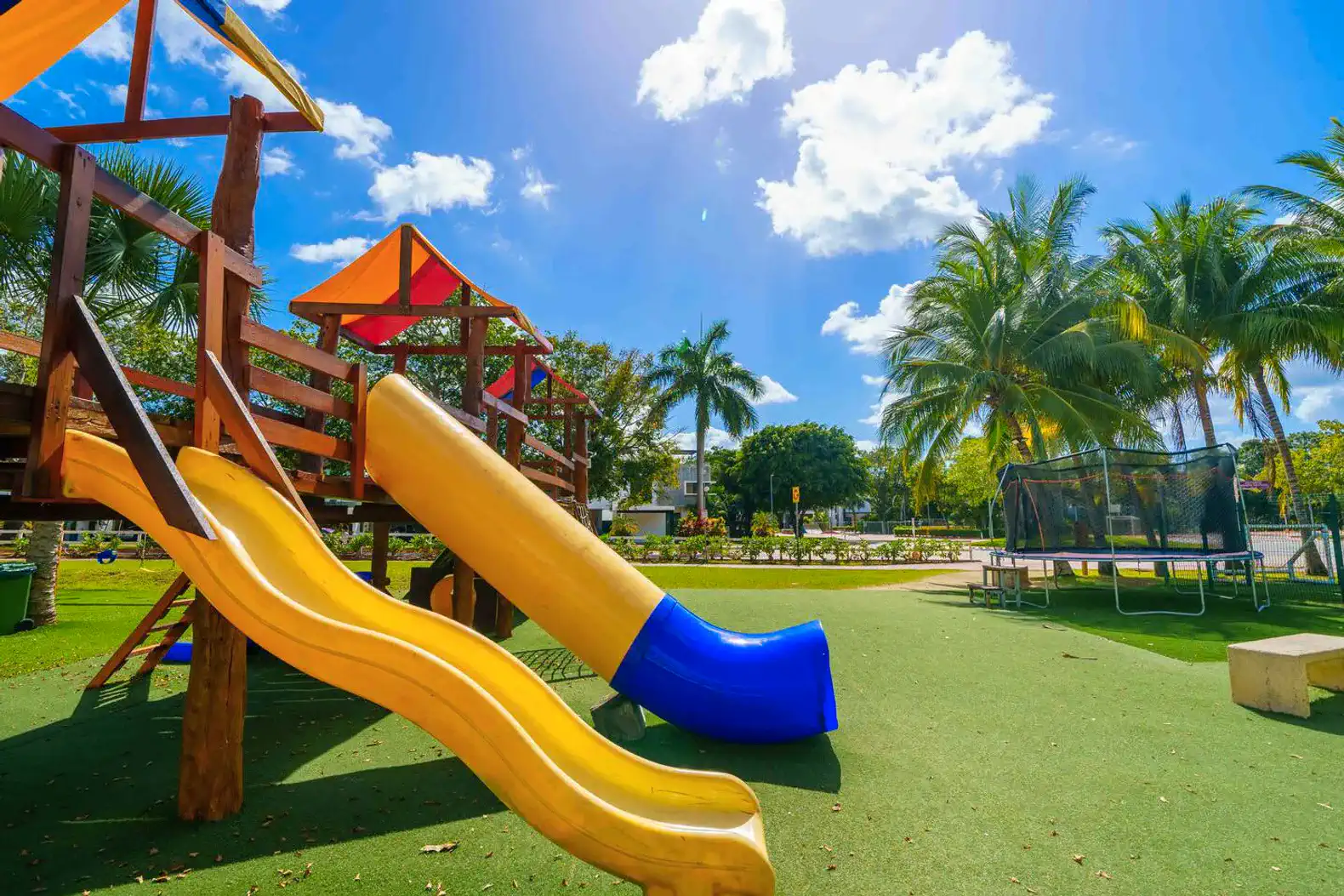 Área de juegos al aire libre con toboganes y trampolín rodeada de palmeras