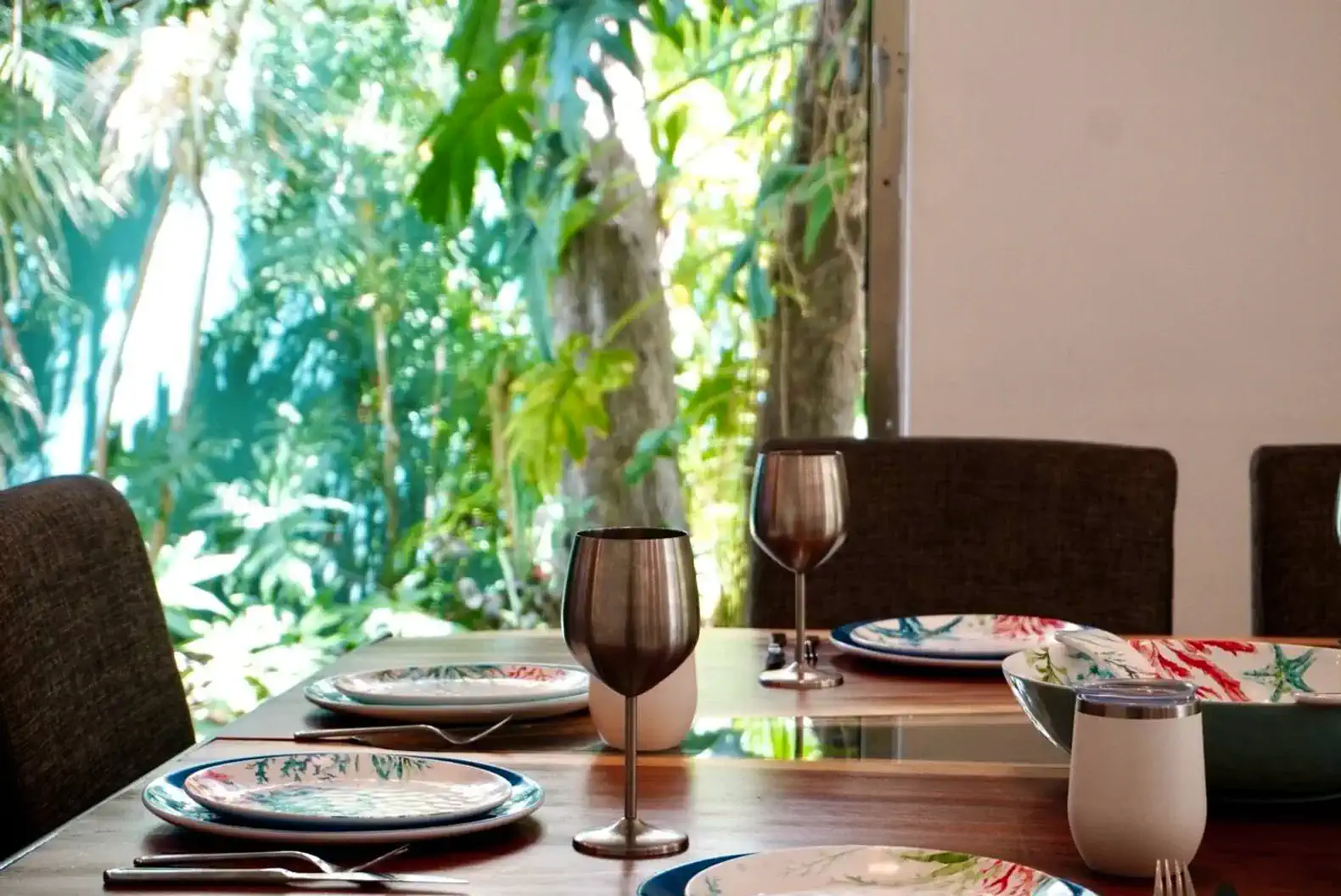Mesa de comedor elegante con platos decorativos y copas, rodeada de vegetación tropical