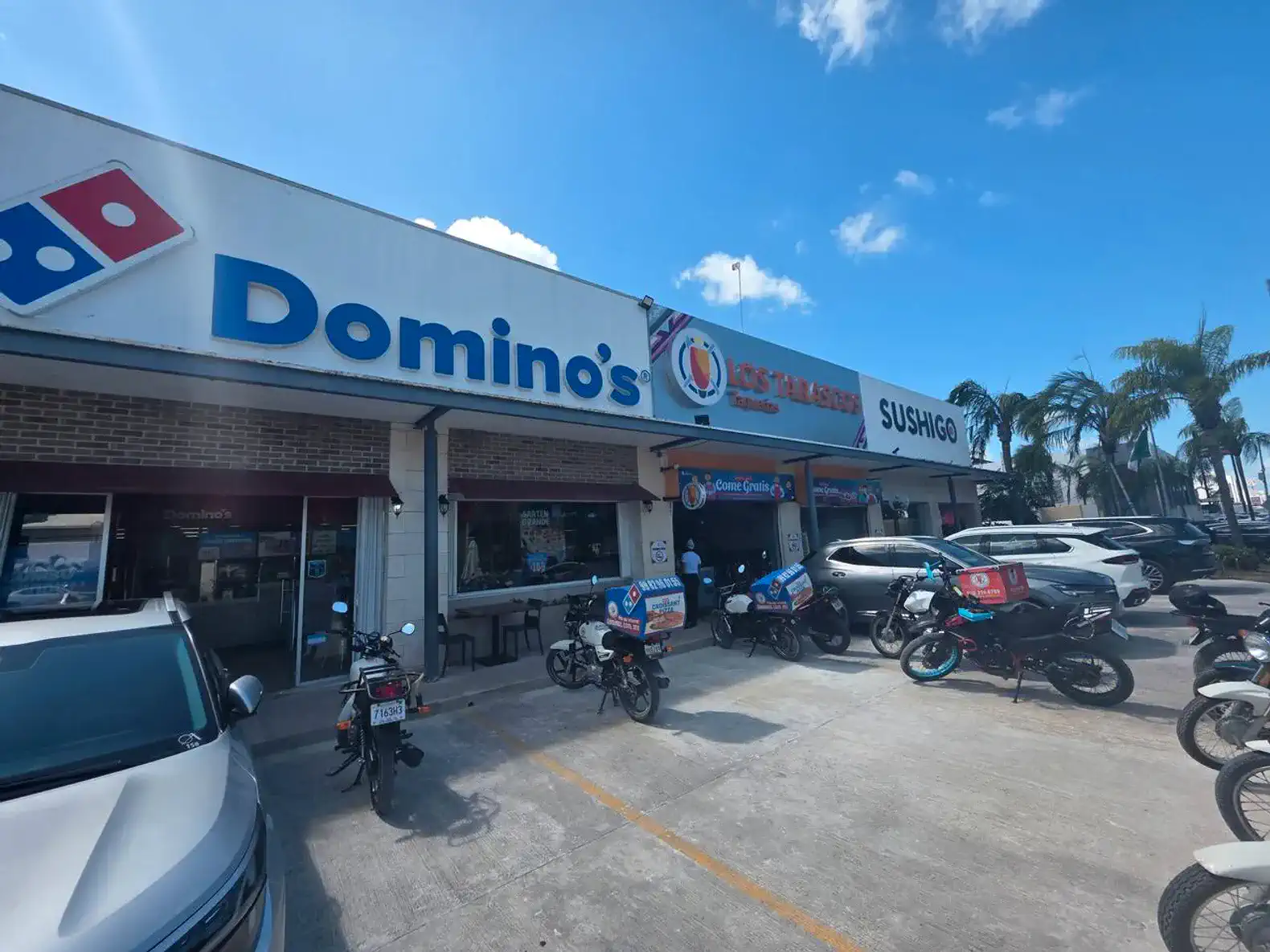 Fachada de un restaurante con letreros de Domino's y Sushi Go, motos estacionadas frente a la entrada
