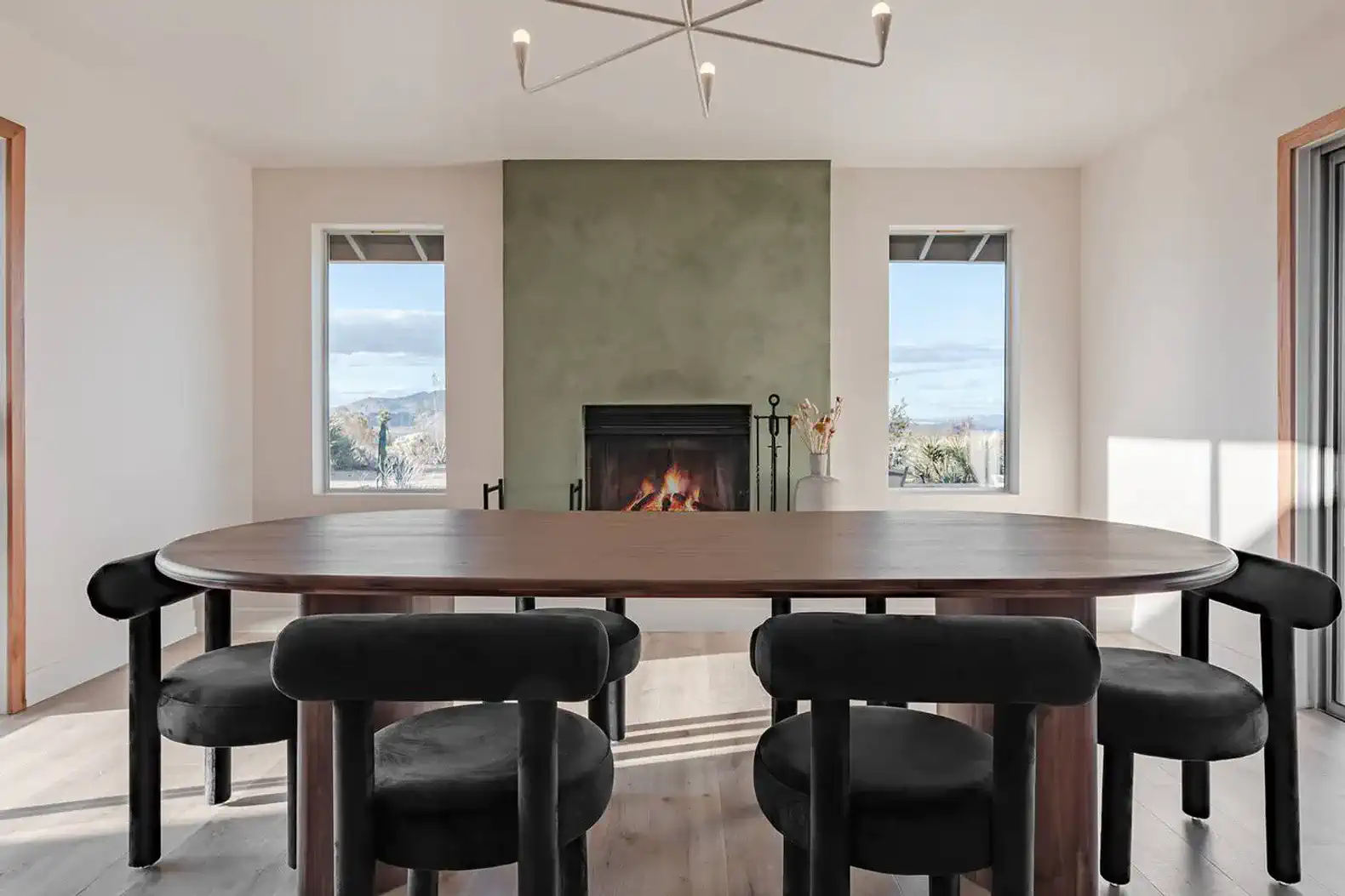Modern dining area with a round table, black chairs, and a fireplace surrounded by large windows