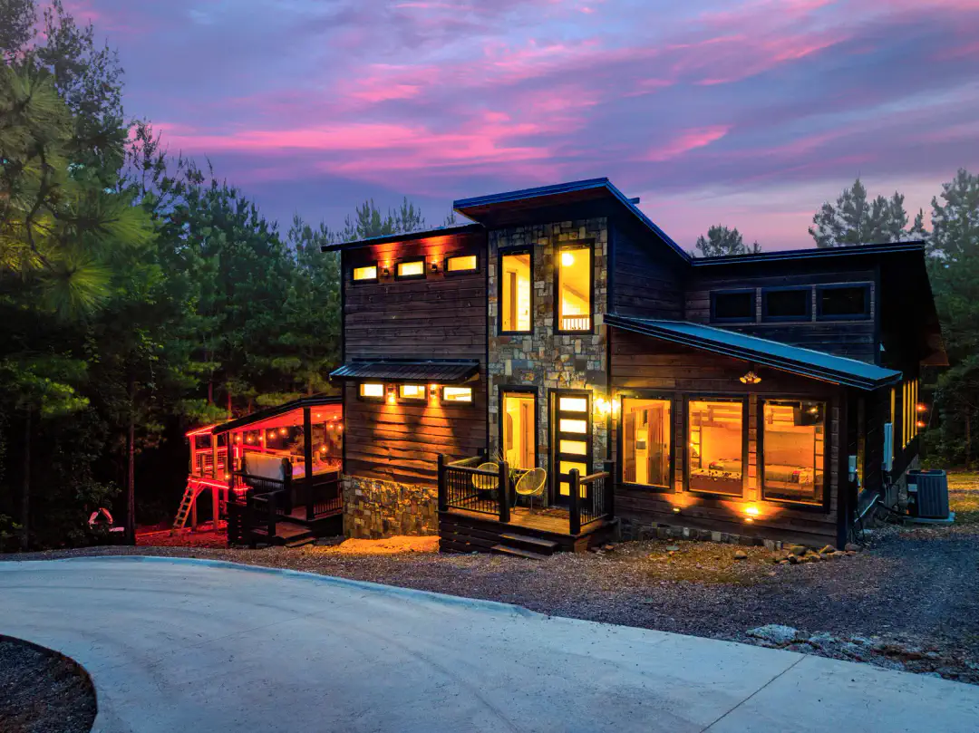 Exterior de cabaña moderna con iluminación cálida y un hermoso atardecer de fondo