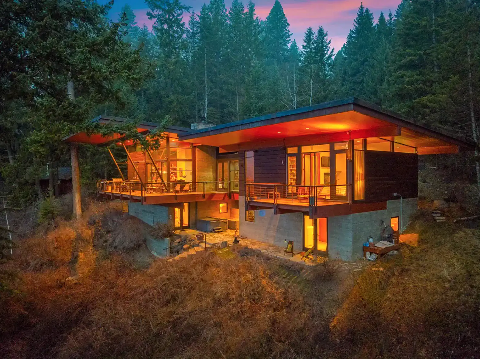 Modern cabin exterior surrounded by trees with warm lights glowing from large windows