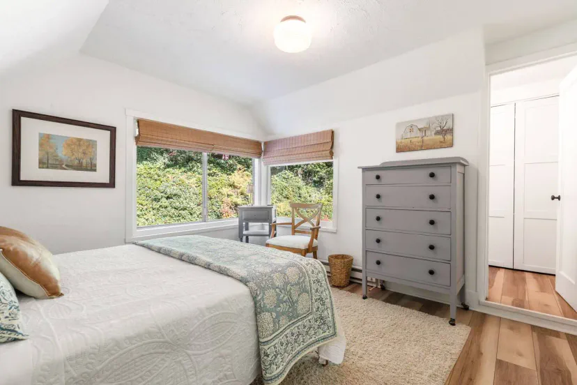 Cozy bedroom with a queen bed, light-colored decor, large windows, and a stylish gray dresser