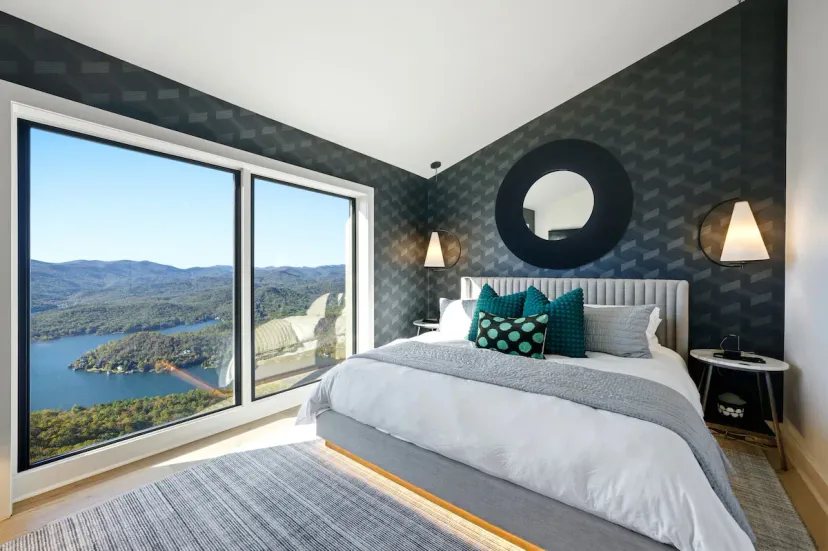 Elegante dormitorio con pared de acento en azul intenso, espejo redondo y ventanas de piso a techo enmarcando vistas de las Montañas Blue Ridge y el Lago Burton — 1 cama king en Tiger, GA.