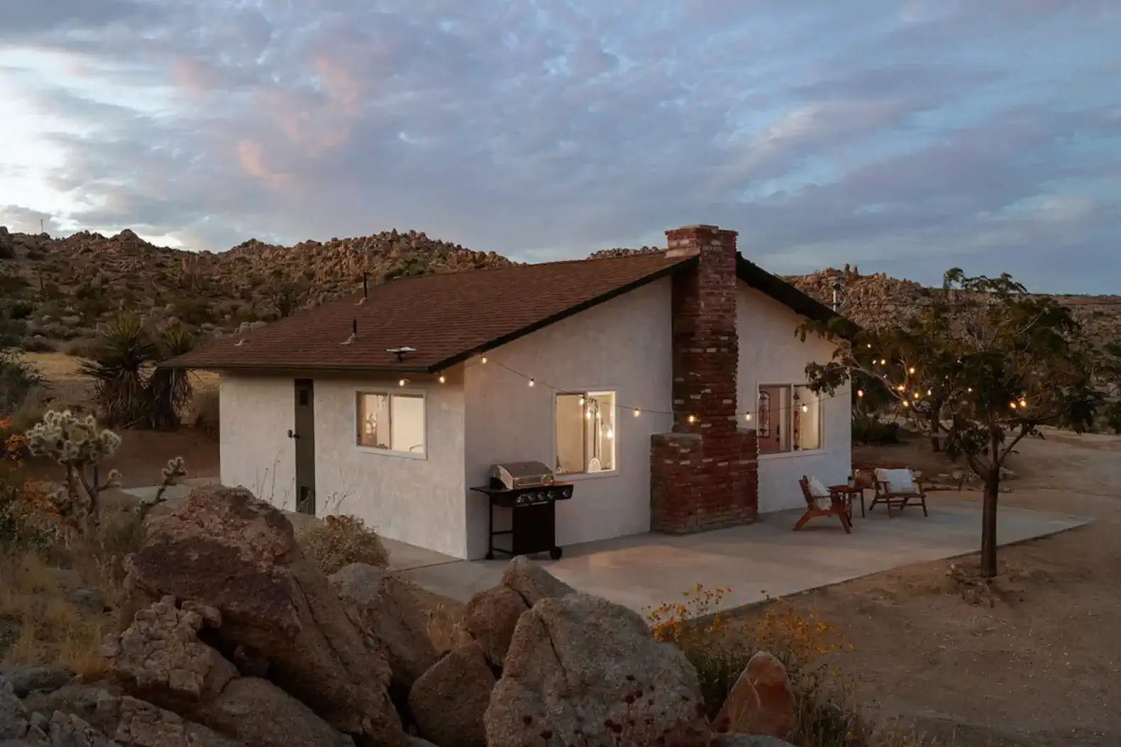 Charming cabin exterior with a stone patio and grill surrounded by boulders and desert landscape